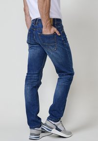 Jeans de mezclilla en azul oscuro con costuras claras, de corte slim, y con bolsillos traseros. Combinados con zapatillas grises con suela blanca.