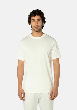 Man met krullend donker haar, gekleed in een effen wit t-shirt met korte mouwen en witte broek, staande tegen een lichte achtergrond.