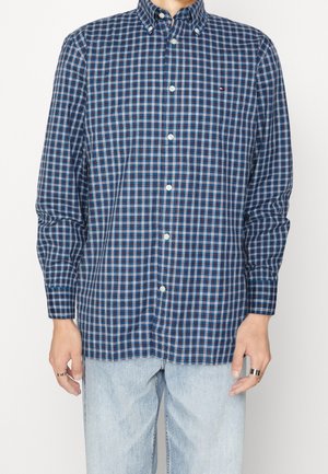 Mężczyzna noszący niebieską, białą i czerwoną koszulę w kratę z długim rękawem na guziki, jasnoniebieskie dżinsy oraz srebrne pierścienie na obu dłoniach.