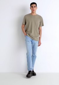 Camiseta verde oliva de manga corta, corte relajado. Jeans azul claro, corte recto. Zapatos negros con suelas gruesas. De pie contra un fondo blanco.