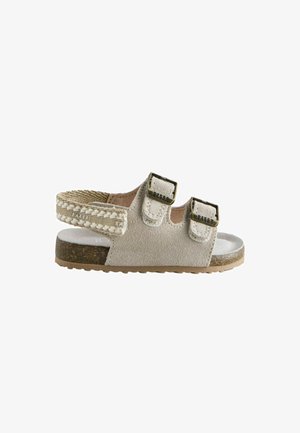 Børnesandal i beige ruskind med to justerbare spænder og vævet bagrem, korkfodbed og gummisål, set fra siden.