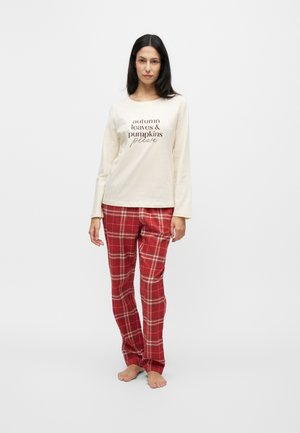 Donna in piedi scalza con maglia a maniche lunghe color crema con scritta autunnale e pantaloni del pigiama a quadri rossi su sfondo bianco semplice.