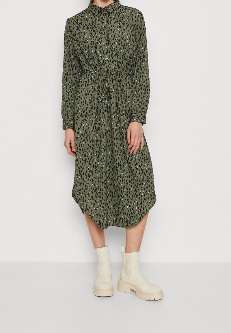 Femme portant une robe longue olive à manches longues avec des taches léopard noires et blanches et des bottines crème, debout devant un fond uni.