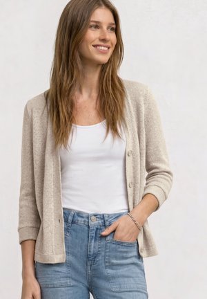 Mujer joven con el pelo largo sonriente, lleva un cárdigan beige sobre una camiseta sin mangas blanca y vaqueros azules, mano en el bolsillo, con un fondo liso.