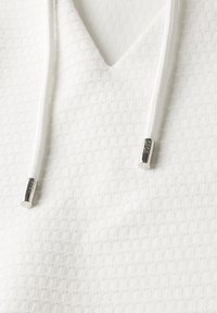 Haut à capuche texturé blanc avec un design en V, comportant des cordons de serrage avec des extrémités métalliques. Le tissu présente un motif en grille pour un ajout de détail.
