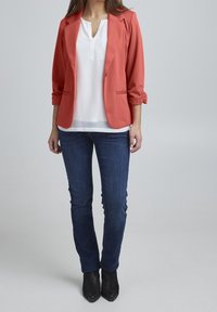 Blazer corail à manches retroussées, associé à un chemisier blanc et un jean skinny bleu foncé, complété par des bottines noires.