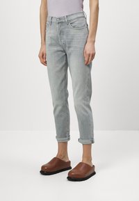Jeans gris clair avec une coupe droite, des poignets retroussés, cinq poches et une fermeture à bouton, associés à des chaussures marron à enfiler avec des semelles texturées.