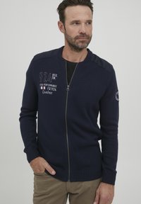 Chalatas su užtrauktuku jūrine mėlyna spalva, su giriamaisiais rankogaliais ir apatine dalimi, ant krūtinės ir pečių detalėmis išsiuvinėtu tekstu, pagamintas iš mezginio.