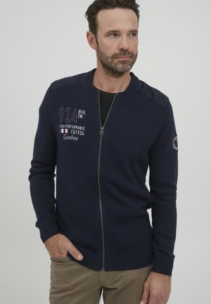 Tengerészkék zipzáras kardigán bordázott mandzsettákkal és alsó résszel, hímzett szöveggel a mellrészen és vállrészletekkel, kötött anyagból készült.