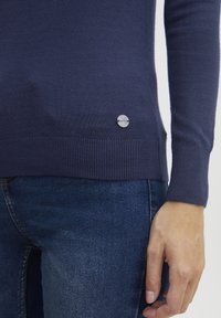 Mörkblå tröja i mjukt tyg med ribbade ärmslut och fåll. Har en metallisk logotypdetalj på den nedre vänstra sidan. Bärs med mörk denim.