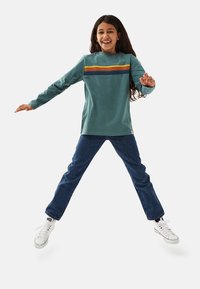 Langärmliges türkisfarbenes Shirt mit horizontalen orangen und marineblauen Streifen, kombiniert mit blauen Jeans und weißen Turnschuhen. Das Modell ist in einer dynamischen Pose.