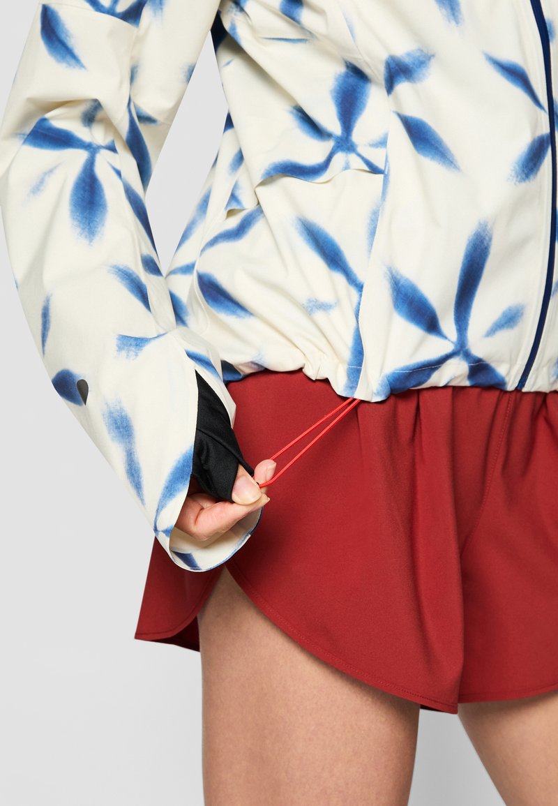 Weißer Jacke mit blauem Blumenmuster, verstellbarem Taillenbund und schwarzen Innenmanschetten, kombiniert mit roten Sportshorts.