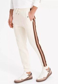 Joggers crema con rayas laterales marrones, cinturilla elástica, cordón y bolsillos delanteros, combinados con zapatillas blancas y marrones.
