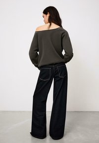 Sötétszürke váll nélküli pulóver ezüst szegecsekkel a nyakrésznél, széles szárú sötét farmernadrággal, hátul zsebekkel.