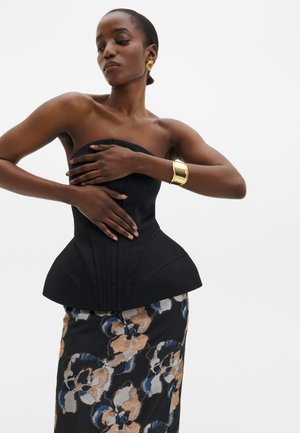 Femme portant un corset noir sans bretelles et une jupe fleurie pose, les bras croisés, accessoirisée avec des boucles d'oreilles dorées et un large bracelet en or.