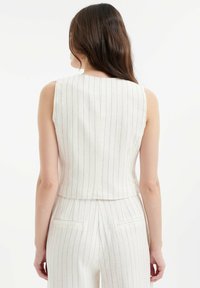 Mouwloos, crèmekleurig stofshirt met verticale marineblauwe strepen. Heeft een aangesloten, cropped ontwerp en subtiele details aan de achterkant.