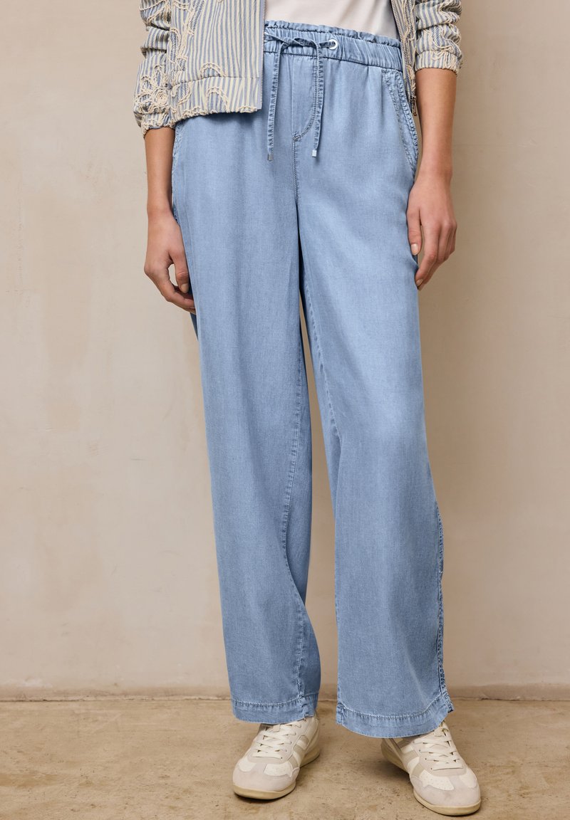 Personne portant un pantalon en jean bleu clair à jambes larges avec taille à cordon, des baskets blanches, et une veste rayée avec les manches retroussées.