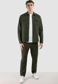 Grøn skjorte med knapper og to brystlommer, parret med matchende grønne bukser og hvide sneakers, sat mod en neutral baggrund.