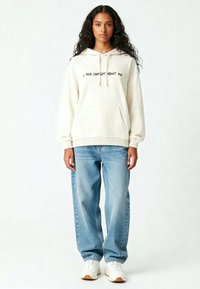 Felpa crema con testo nero "HO PARLATO DI TE A CHAT GPT", tasca frontale, abbinata a jeans baggy azzurri e sneakers bianche.