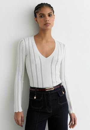 Biely prúžkovaný top s dlhým rukávom a hlbokým výstrihom do V so zvislými pruhmi. Kombinovaný s tmavomodrými džínami s vysokým pásom a hnedým opaskom.