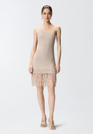 Donna che indossa un abito beige all'uncinetto monospalla con orlo a frange, orecchini a cerchio dorati e sandali beige con tacco alto e dettagli con borchie.