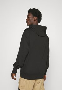 Felpa nera con cappuccio, dal fit rilassato, con polsini e orlo a costine e tasca frontale a marsupio. Il modello indossa pantaloni cargo beige.