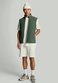Green zip-up vest with a small logo, white textured short-sleeve shirt, white shorts, white beanie, and neutral sneakers.