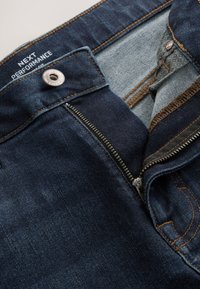 Jeans denim blu scuro con zip e bottone aperti, fodera interna visibile ed etichetta bianca con testo che dice "NEXT PERFORMANCE."