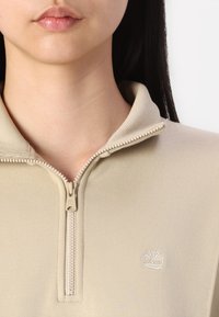 Maglione beige con zip e alto collo, caratterizzato da una texture a coste e un piccolo logo bianco a forma di albero sulla parte sinistra del petto.
