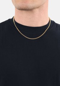 Primo piano di una persona che indossa una camicia nera e una sottile collana d'oro intorno al collo, su uno sfondo semplice.