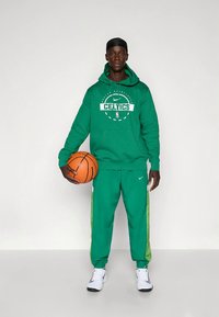 Felpa verde Nike con grafica "CELTICS", pantaloni della tuta verdi abbinati con strisce laterali bianche e scarpe da ginnastica bianche che tengono un pallone da basket.