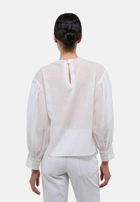 Blouse blanche à manches longues en tissu transparent, avec des manches bouffantes, un bord en dentelle aux poignets et une fermeture en forme de clé au dos. Texture lisse et légère.