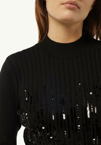 Schwarzer Rippstrickpullover mit hohem Kragen, der vertikale Paillettenakzente aufweist, die in der Größe variieren und einen strukturierten, schimmernden Effekt erzeugen.