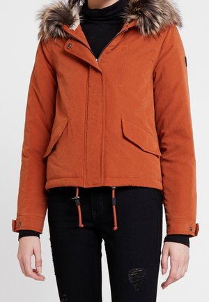 Mujer con chaqueta de invierno color naranja óxido con capucha con borde de piel, suéter de cuello alto negro y jeans negros desgarrados, posando frente a un fondo liso.
