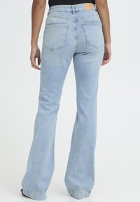 Persoon die lichtblauwe, aansluitende, uitlopende hoge taille jeans draagt en staat tegen een effen witte achtergrond, gezien van achteren.