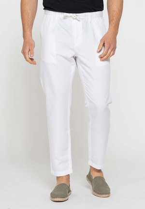 Uomo che indossa pantaloni bianchi con coulisse e scarpe slip-on verde oliva su sfondo neutro.
