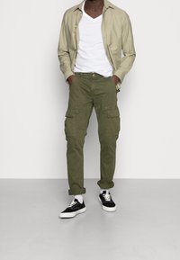 Olivgröna cargo-byxor med flera sidofickor, bärs med en vit V-ringad tröja och en ljus olivgrön skjorta med knappar, kombineras med svarta sneakers.