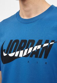 Približek modre majice z velikim črno-belim besedilom "JORDAN" in logotipom Nike swoosh čez prsni koš.