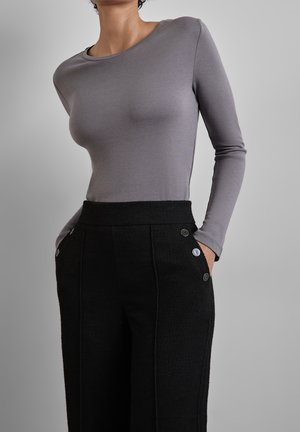Frau mit kurzen lockigen Haaren, die ein eng anliegendes graues Langarmshirt und hoch taillierte schwarze Hose mit Knöpfen trägt, die Hände in den Taschen.