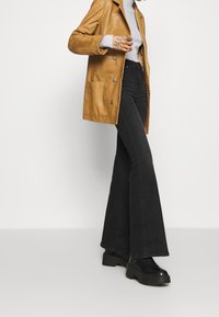 Henkilö, jolla on päällään vaaleanruskea nahkatakki, vaaleanharmaa ribattua villapaitaa, mustat levenevät farkut ja mustat korokepohjaiset saappaat, seisoo valkoista taustaa vasten.