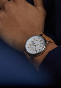 Polso che indossa un orologio Timex d'argento con quadrante bianco, indicatori dorati e cinturino in pelle color cuoio, che mostra l'ora 4:45 e la data lunedì 29.