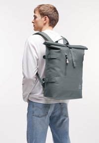 Mochila enrollable gris hecha de material impermeable. Cuenta con correas de hombro ajustables, hebillas laterales y un bolsillo frontal con cremallera.