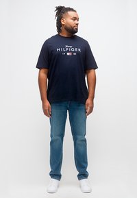 Άνδρας με dreadlocks που φοράει μπλε μπλουζάκι Tommy Hilfiger, μπλε τζιν και λευκά αθλητικά παπούτσια, στέκεται και κοιτάζει δεξιά σε λευκό φόντο.