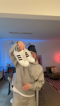 Inimene halli kapuutsiga ja musta mütsiga, hoides valgeid tossusid mustade triipudega moodsas elutoas, kus on värviline valgustus.