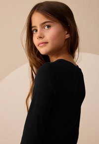 Top negro de manga larga con textura acanalada y cuello redondeado, usado por un niño. Diseño ajustado y textura suave. Fondo en tono beige claro.