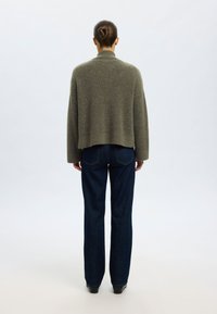 Prevelik pulover olivno zelene barve z visokim ovratnikom, mehkim otipom in spuščenimi rameni, kombiniran z temno modrimi kavbojkami s pravimi hlačnicami.