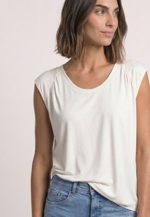 Kvinde med skulderlangt brunt hår iført en ærmeløs hvid top og blå jeans, der ser til siden med et neutralt udtryk.