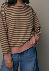 Persona che indossa una felpa a righe orizzontali rosa e verde oliva con jeans di denim blu chiaro, una mano in tasca, su uno sfondo semplice.