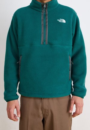 Person mit einer petrolfarbenen The North Face Fleecejacke mit halbem Reißverschluss und braunen Hosen, von den Schultern bis zu den Oberschenkeln vor einem schlichten Hintergrund gezeigt.
