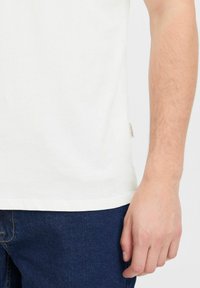 Fehér pamut t-shirt, kerek nyakkivágással, rövid ujjakkal és egy kis címke részlettel az alján, kék farmerral párosítva.